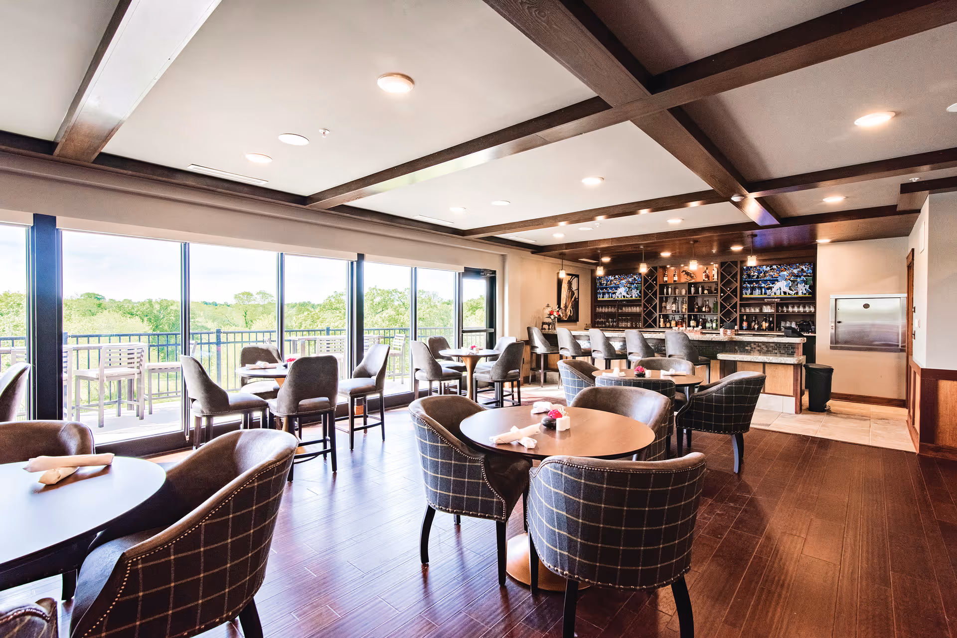 A bright and spacious dining area with large windows offering a view of greenery outside. The room features several round tables with cushioned chairs arranged around them. In the background, there is a bar area with high stools, shelves stocked with bottles, and two televisions mounted on the wall. The ceiling has wooden beams and recessed lighting, and the floor is made of dark wood.