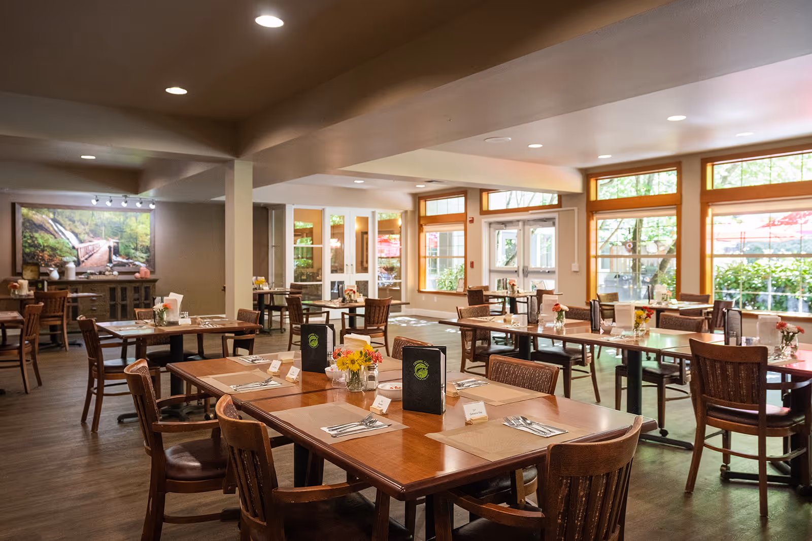 A bright and spacious dining room with multiple wooden tables and chairs arranged neatly. Each table is set with placemats, utensils, menus, and small flower vases. Large windows allow natural light to fill the room, and a large framed photograph of a forest scene hangs on one wall.