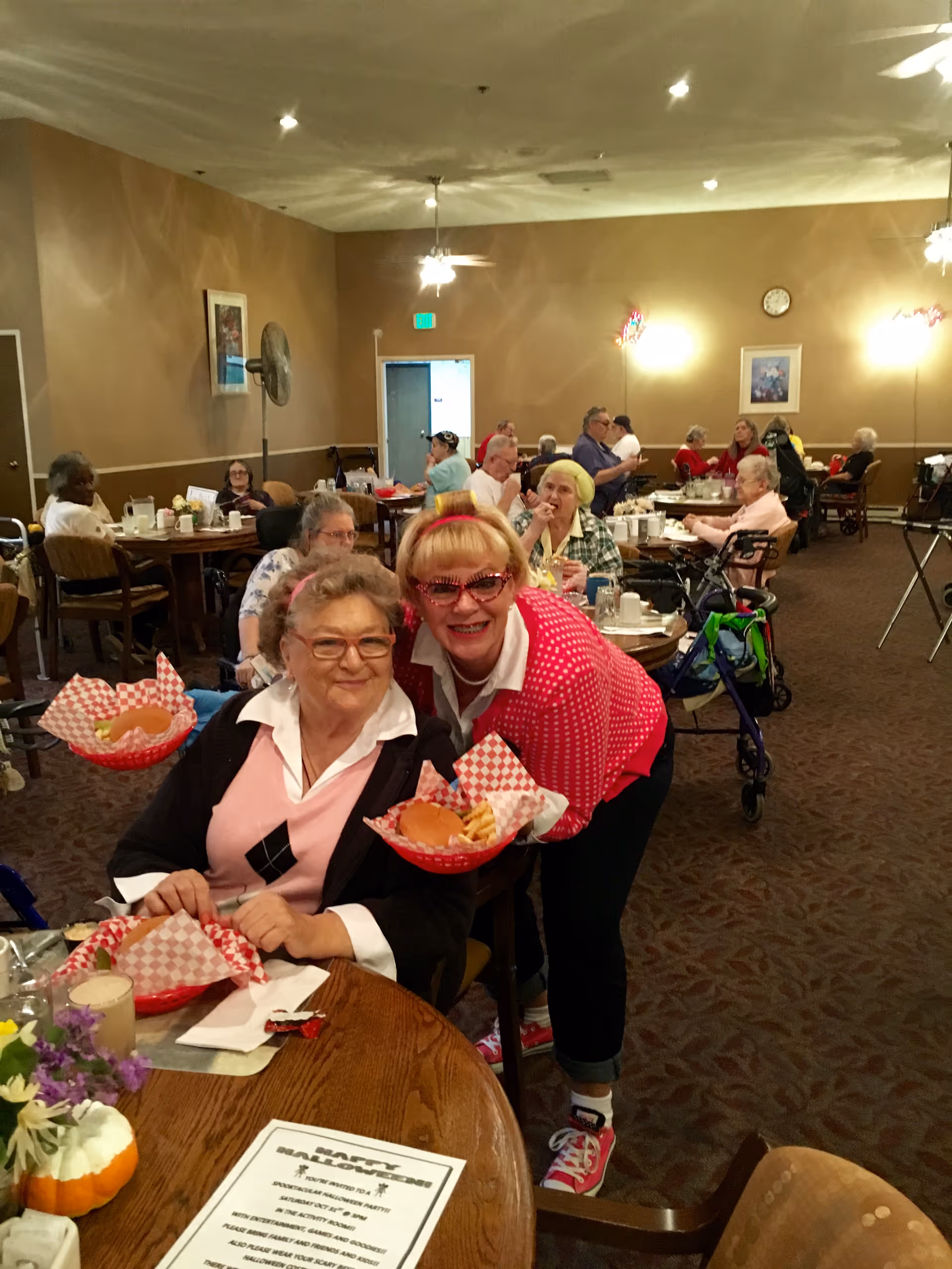 A dining room in Charlton Place with elderly residents seated at tables enjoying meals. Two women are in the foreground, one seated and one standing behind her, both smiling and holding baskets with burgers and fries. Other residents are seated at tables in the background, some using walkers. The room has warm lighting, beige walls, and framed pictures on the walls.