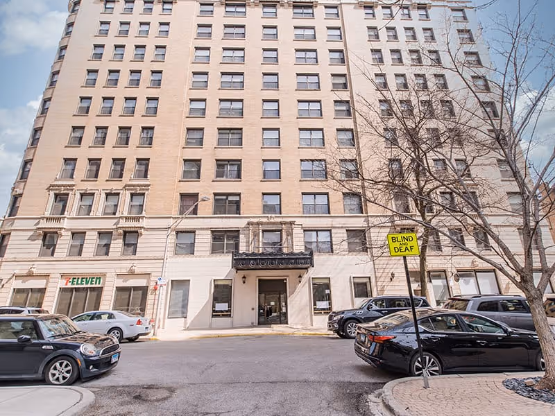 Front exterior of a multi-story beige apartment building with a covered entrance, cars parked in front, and a 7-Eleven storefront at street level.