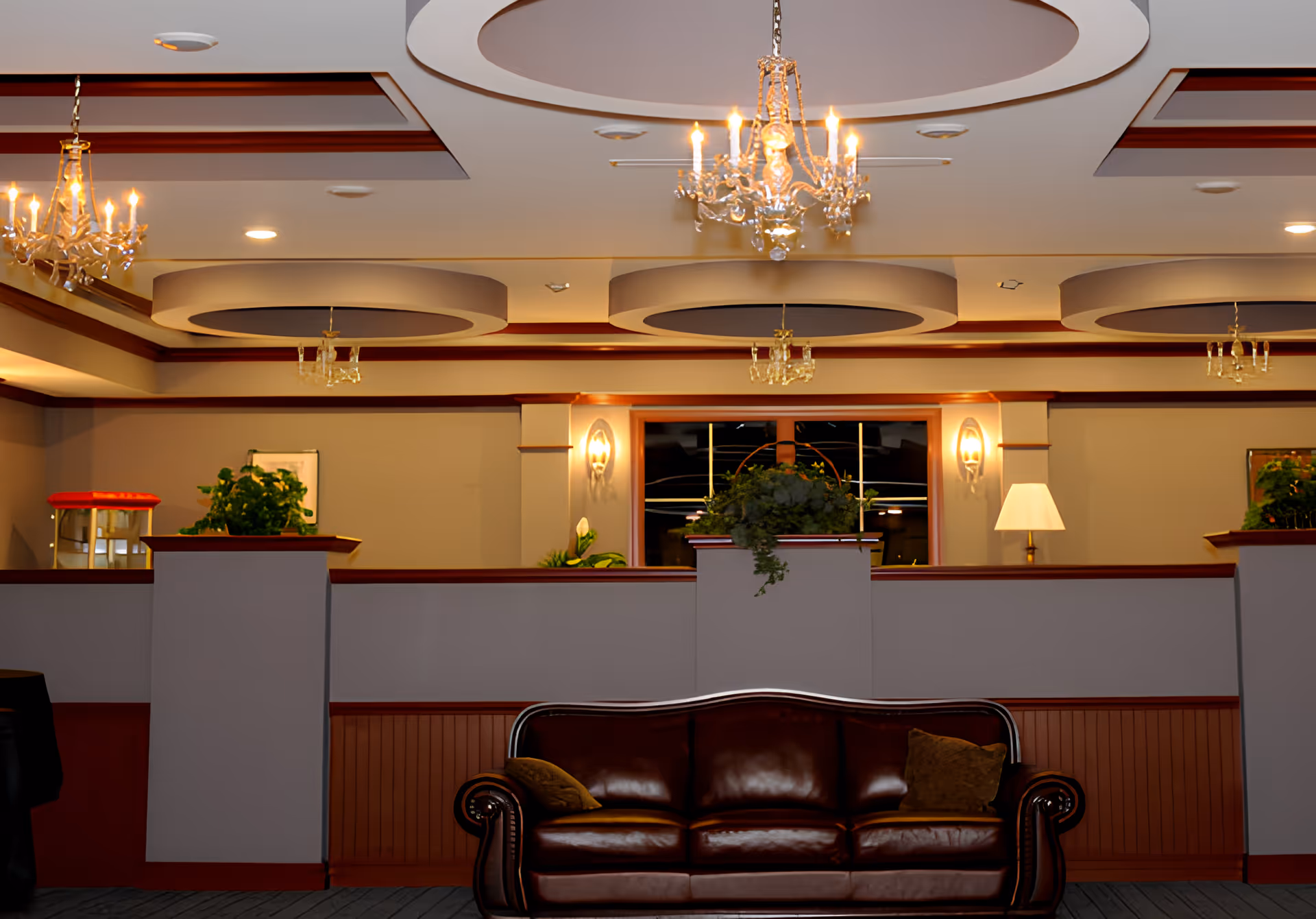 Interior view of a senior living facility lounge area with a dark brown leather sofa in the foreground, two pillows on the sofa, decorative plants on a half wall behind it, and multiple chandeliers hanging from a ceiling with circular recessed designs.