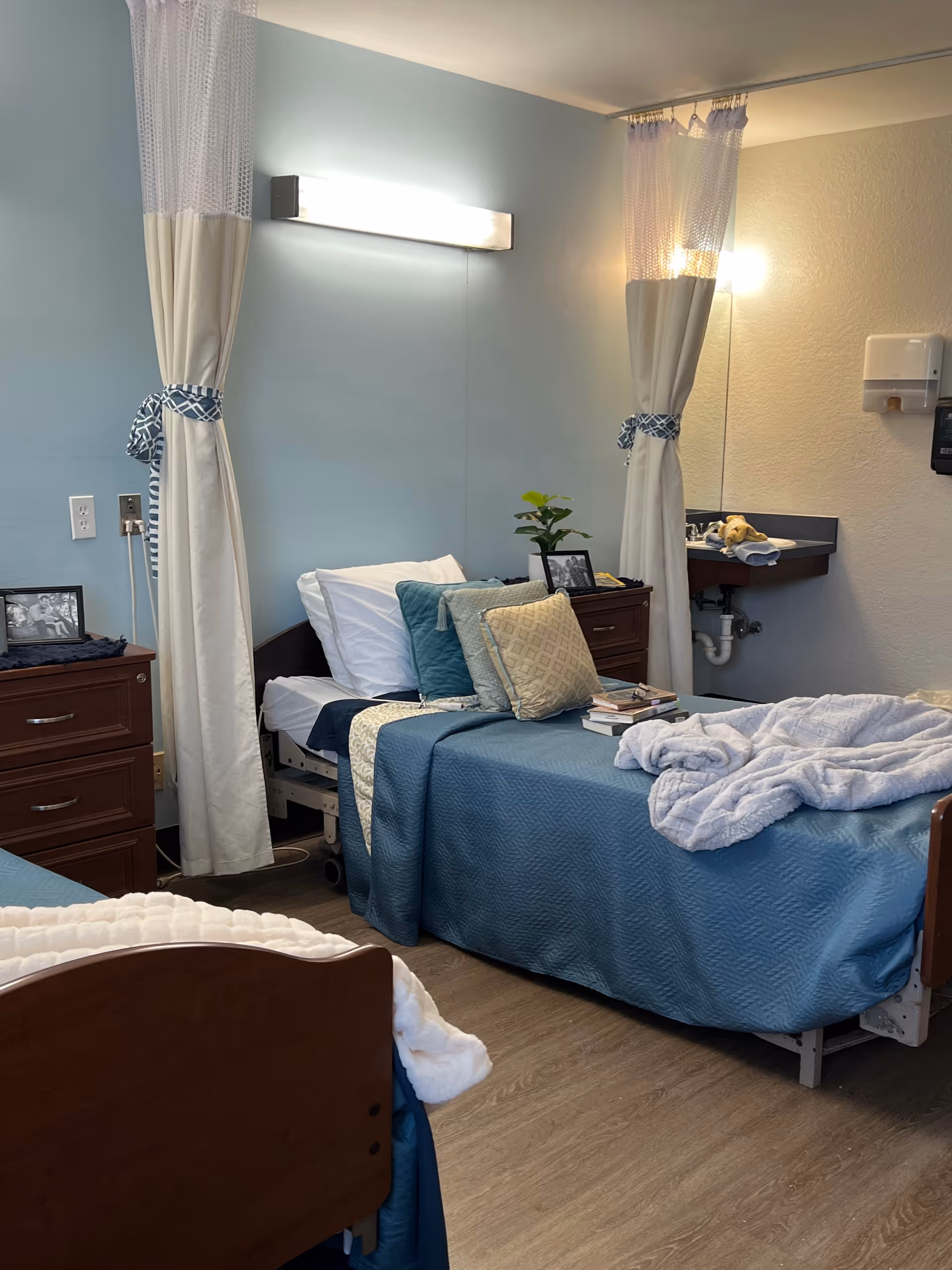 A healthcare facility room with two hospital beds, each made with blue bedding and multiple pillows. There are privacy curtains partially drawn around each bed. A wooden nightstand with framed photos and a small plant is positioned between the beds. A wall-mounted light fixture illuminates the room, and a small sink with a paper towel dispenser is visible on the right wall.