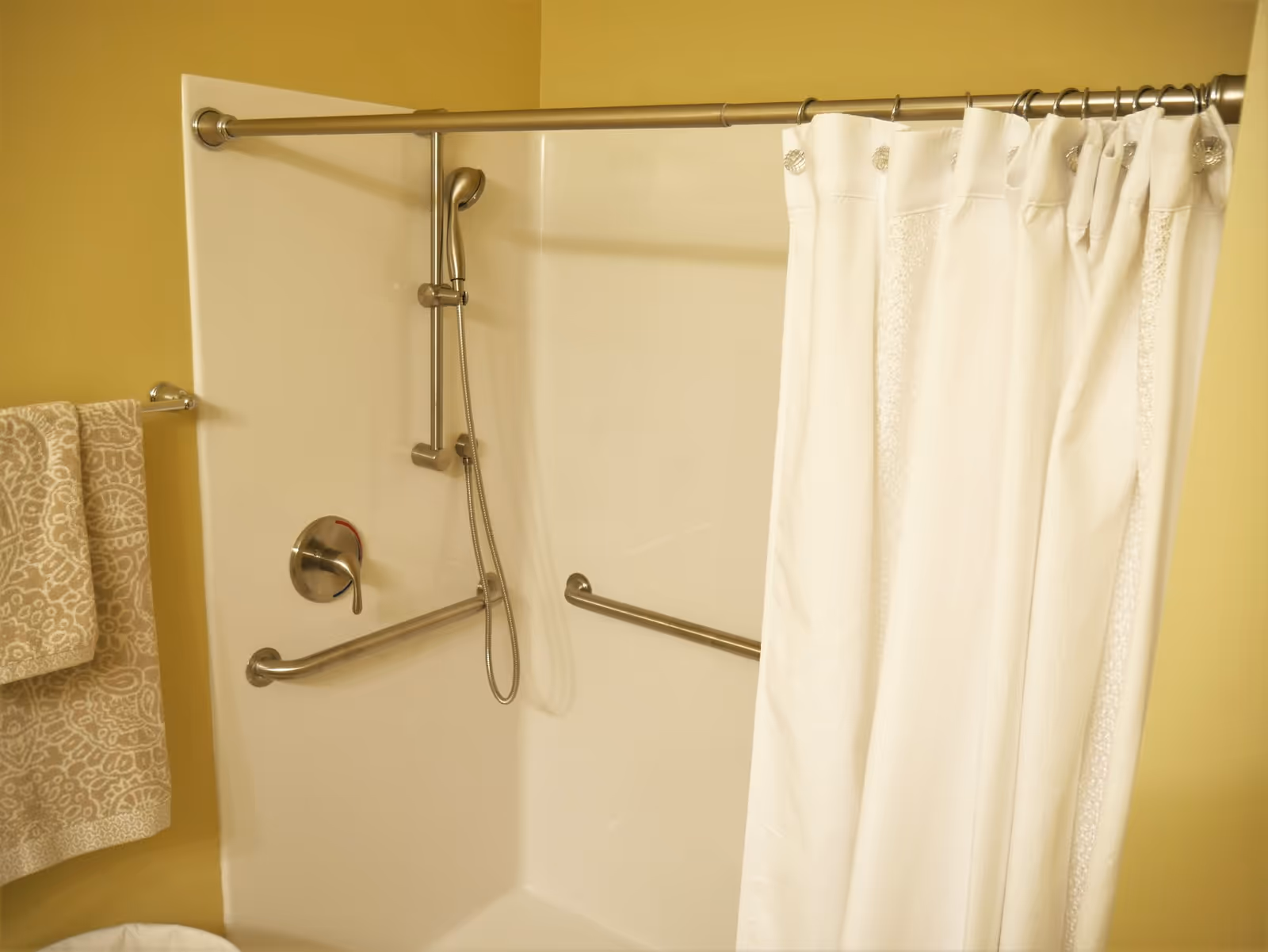 A bathtub/shower with a white curtain, handheld showerhead, and stainless steel grab bars.