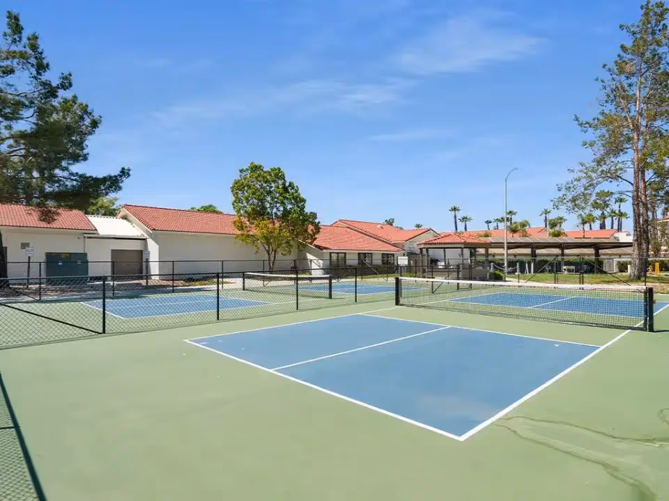 Outdoor view of a pickleball court with multiple playing areas surrounded by a black chain-link fence. In the background, there are single-story buildings with red tile roofs, trees, and a clear blue sky.