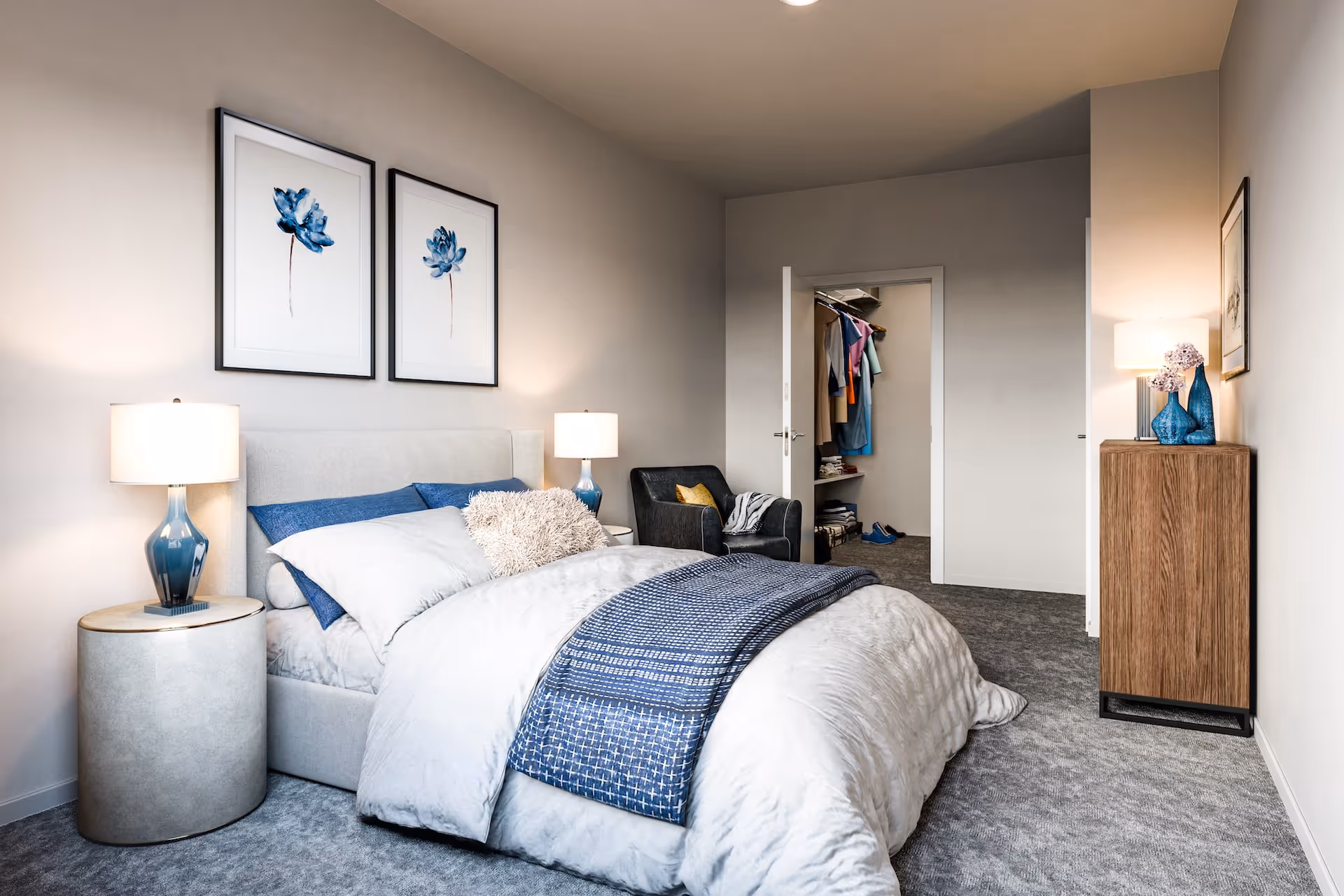 A neatly staged bedroom with a bed, bedside lamps and tables, framed artwork, an armchair, dresser, and an open closet.