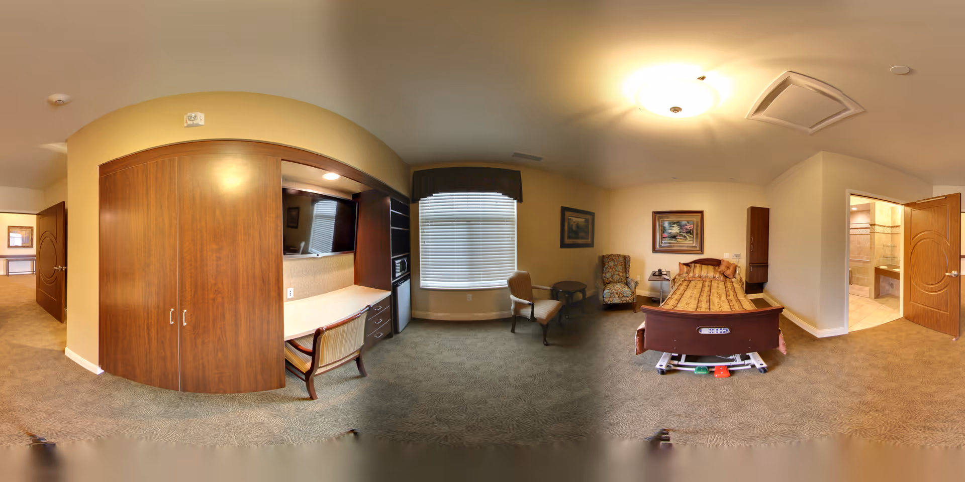 A senior living facility room with a single adjustable bed, a patterned armchair, a wooden chair, a small round table, a large window with blinds, a built-in desk with a chair, and a kitchenette area with a microwave and refrigerator. The room has beige walls, carpeted floor, and a bathroom visible through an open door.