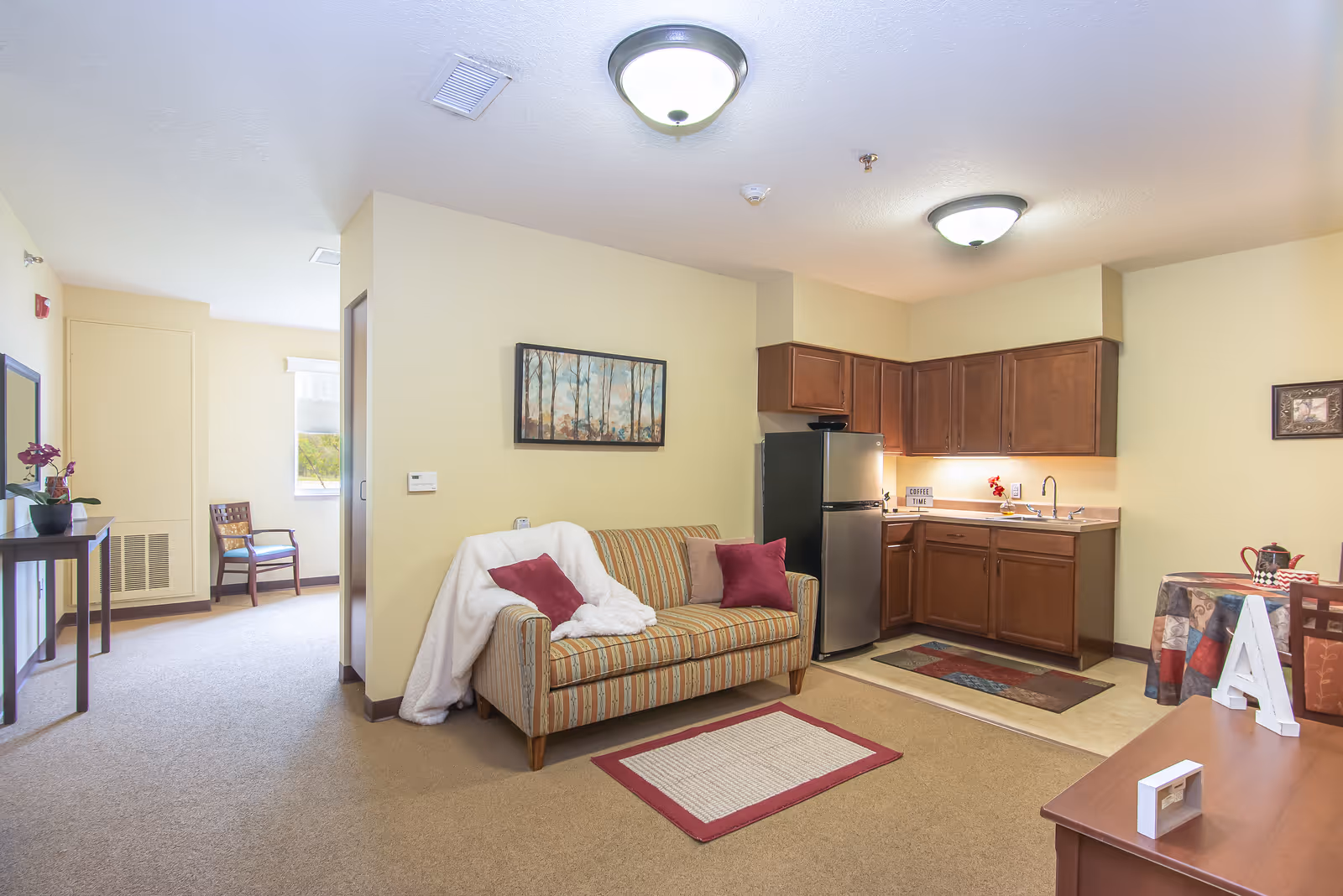 Studio living area with a striped sofa, throw pillows and blanket, a small kitchenette with wood cabinets and stainless refrigerator, and a dining table.