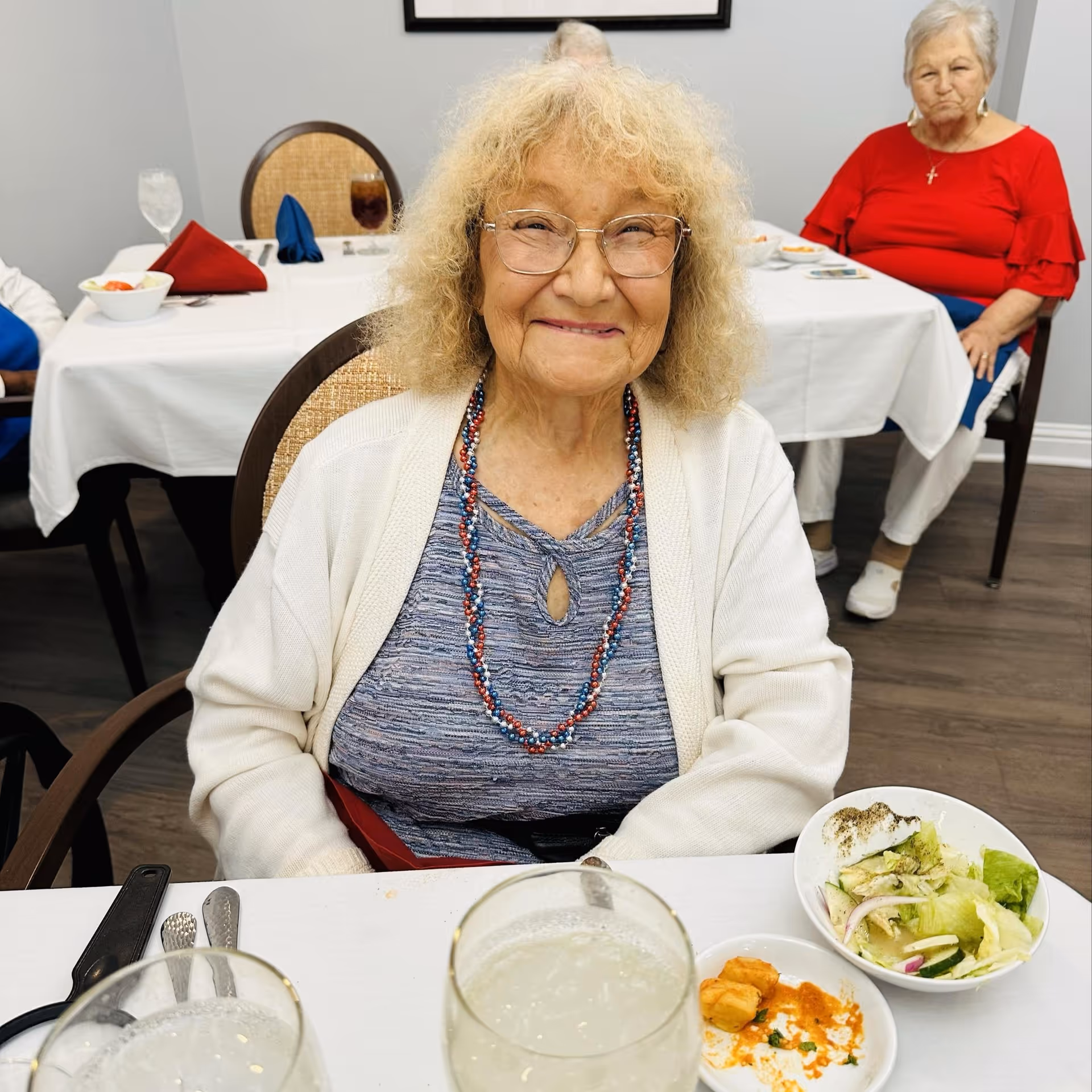 An elderly woman with curly blonde hair, glasses, and a beaded necklace is smiling while sitting at a dining table with plates of salad and food in front of her. In the background, another elderly woman wearing a red top is seated at a table with white tablecloths and folded napkins.