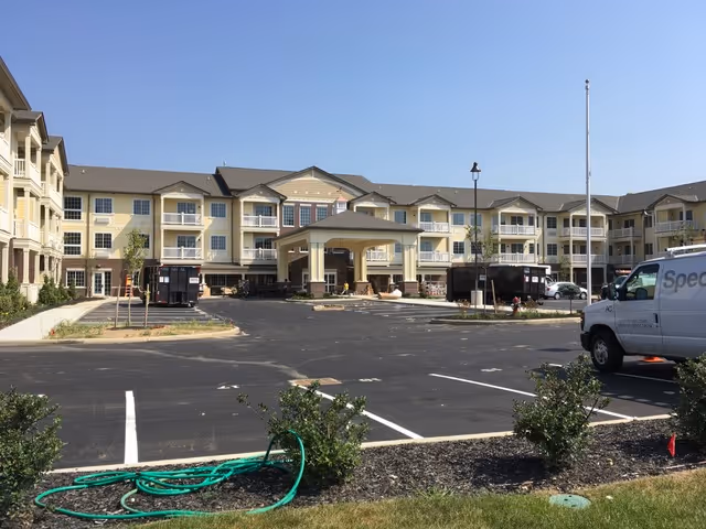 Exterior view of a three-story senior living facility building with beige siding and brick accents. The building has multiple balconies and a covered entrance. In front of the building is a large parking lot with some vehicles and landscaping including small bushes and a green garden hose on the ground.