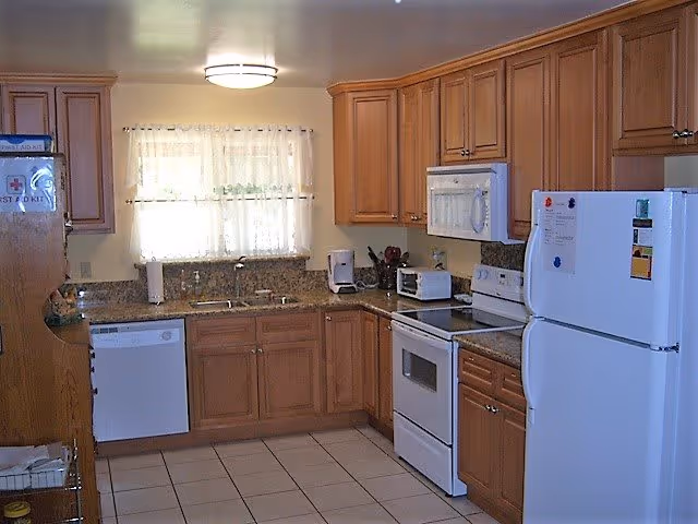 A clean kitchen with wooden cabinets, a white refrigerator, white stove with oven, white microwave, dishwasher, and a coffee maker on the granite countertop. There is a window with sheer curtains above the sink letting in natural light.
