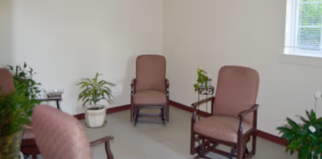 A small sitting area with four cushioned wooden chairs arranged in a circle. There are several potted plants placed around the room, and a window with blinds lets in natural light. The walls are plain and light-colored.