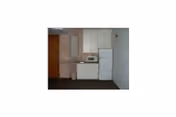 Small kitchenette area with white cabinets, a microwave on the countertop, a white refrigerator, and a trash can next to the counter. The walls are plain and the floor is dark-colored.