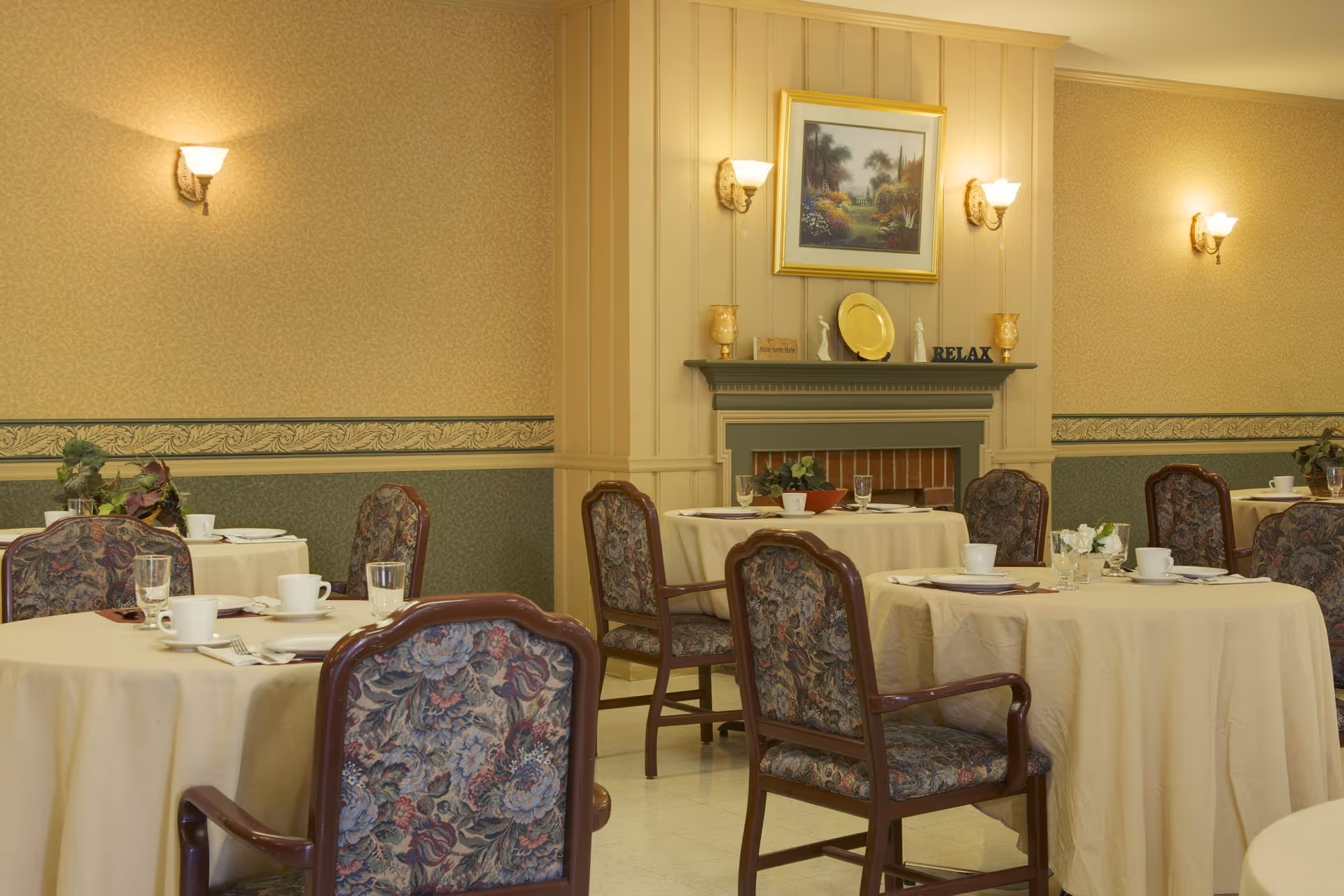 A dining room with round tables covered in beige tablecloths, each set with white cups, glasses, and silverware. The chairs have wooden frames with floral-patterned upholstery. The room features a decorative fireplace with a painting above it and wall sconces providing warm lighting.