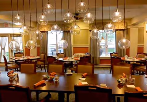 Dining room with multiple tables set with orange napkins, cups, and small decorative items. The room features large windows with curtains and modern hanging globe light fixtures.