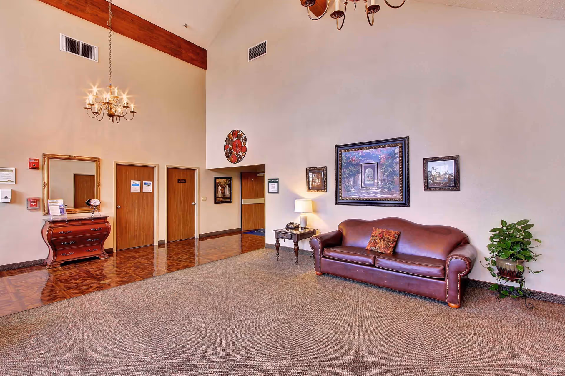 A spacious interior common area with a brown leather sofa, a side table with a lamp and telephone, framed artwork on the walls, a potted plant, a wooden chest of drawers with a mirror above it, and three wooden doors. The floor is carpeted with a polished wooden section near the doors, and a chandelier hangs from the ceiling.