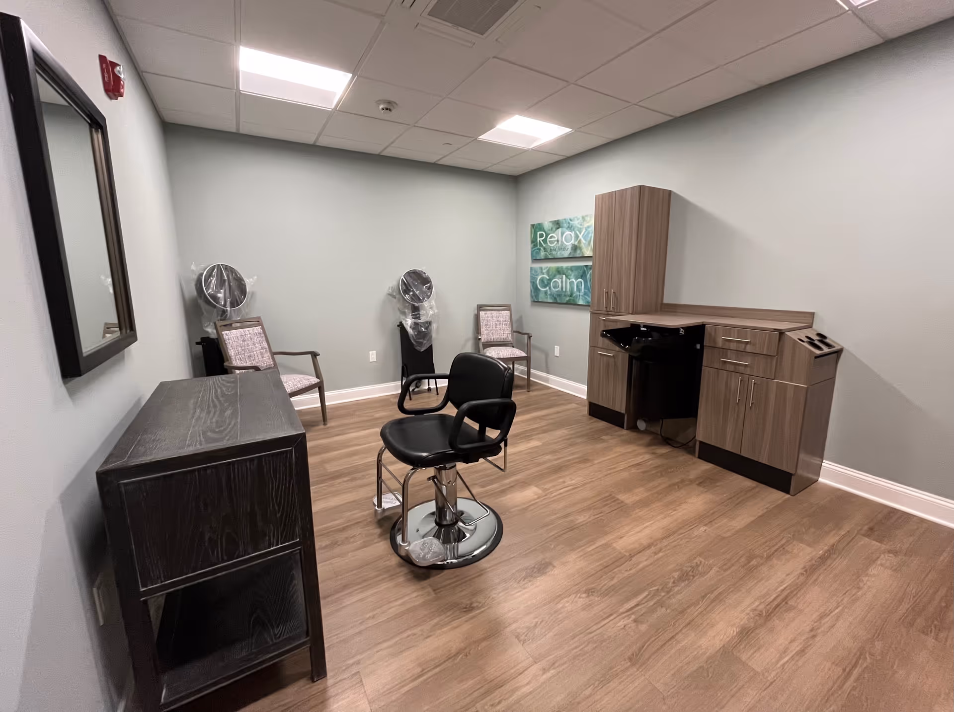 A small salon room with a black salon chair in the center, two chairs with hair dryers covered in plastic against the back wall, a wooden cabinet with drawers and a built-in sink, a black wooden cabinet on the left, and two wall signs that say 'Relax' and 'Calm'. The room has light green walls and wood flooring.