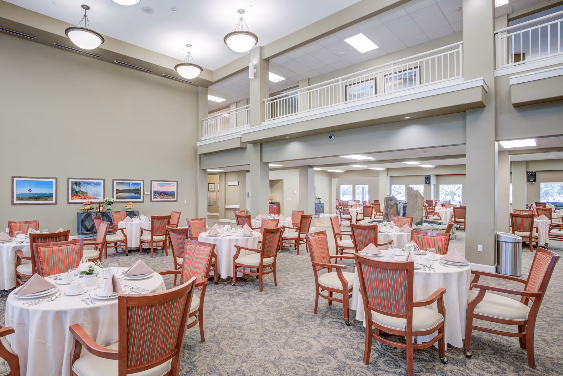 Spacious dining room in a senior living facility with round tables covered in white tablecloths, set with plates, cups, and napkins. The room features high ceilings with hanging light fixtures, carpeted floors, and framed landscape photographs on the wall. There is a second-floor balcony railing overlooking the dining area.
