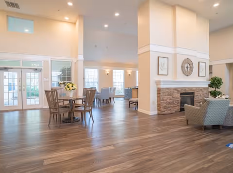 Spacious and well-lit common area in a senior living facility with wooden flooring, a round table with chairs, a stone fireplace with a clock and framed artwork above it, and additional seating near large windows letting in natural light.