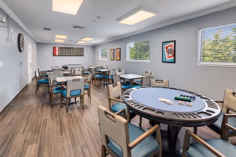 A bright and spacious dining room with multiple tables and chairs arranged neatly. The room has light gray walls, wood flooring, and several windows letting in natural light. There is a round poker-style table in the foreground with poker chips and cards on it. The walls are decorated with framed artwork, and a large bingo board is mounted on the far wall.