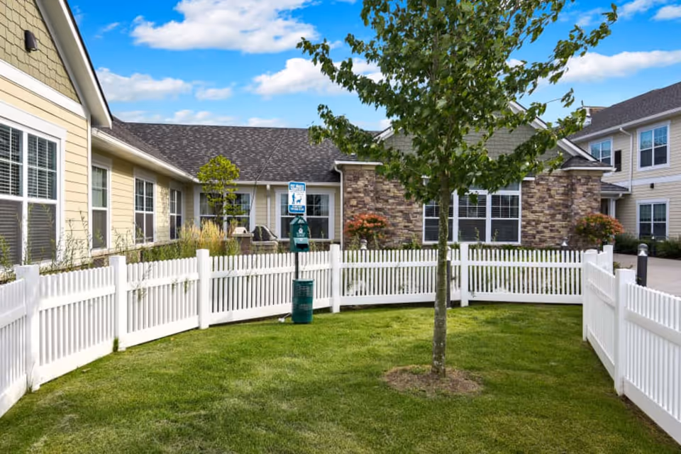 Small fenced courtyard with a young tree and green lawn in front of a residential building with stone and siding.