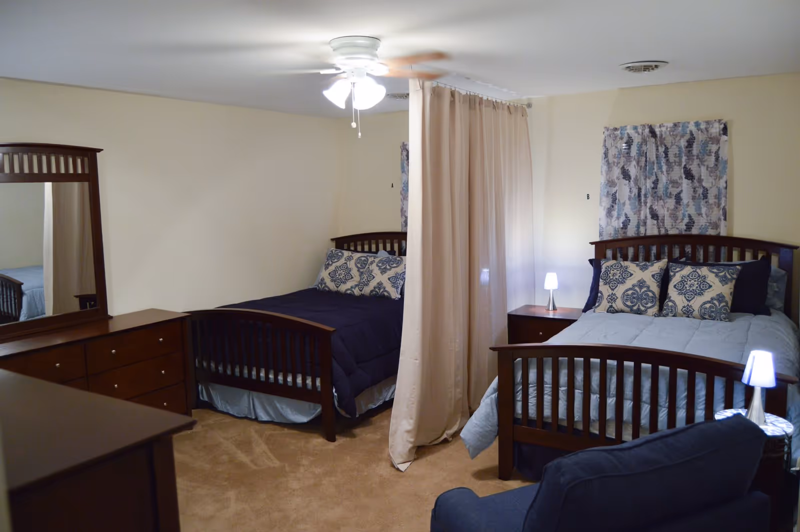 A shared bedroom with two beds separated by a beige curtain. Each bed has a wooden frame and patterned pillows. There are two bedside tables with small lamps, a wooden dresser with a mirror, and a ceiling fan with lights. The room has beige walls and carpeted floor.