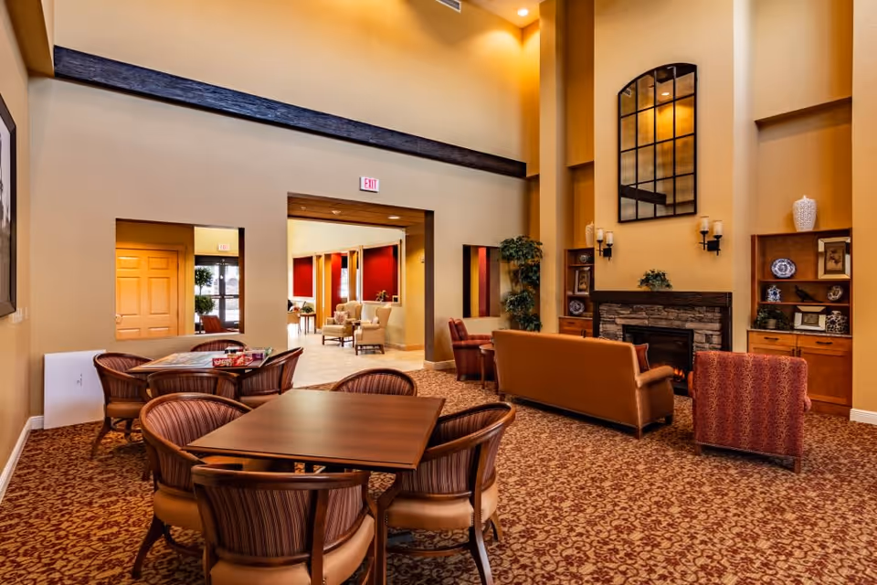 A spacious senior living facility common area with high ceilings, warm yellow walls, and patterned carpet. The room features a seating area with a brown leather sofa, a red patterned armchair, a stone fireplace with decorative shelves on either side, and a large window-like mirror above the fireplace. In the foreground, there are two wooden tables surrounded by striped cushioned chairs. The room opens into another area with additional seating and red accent walls.