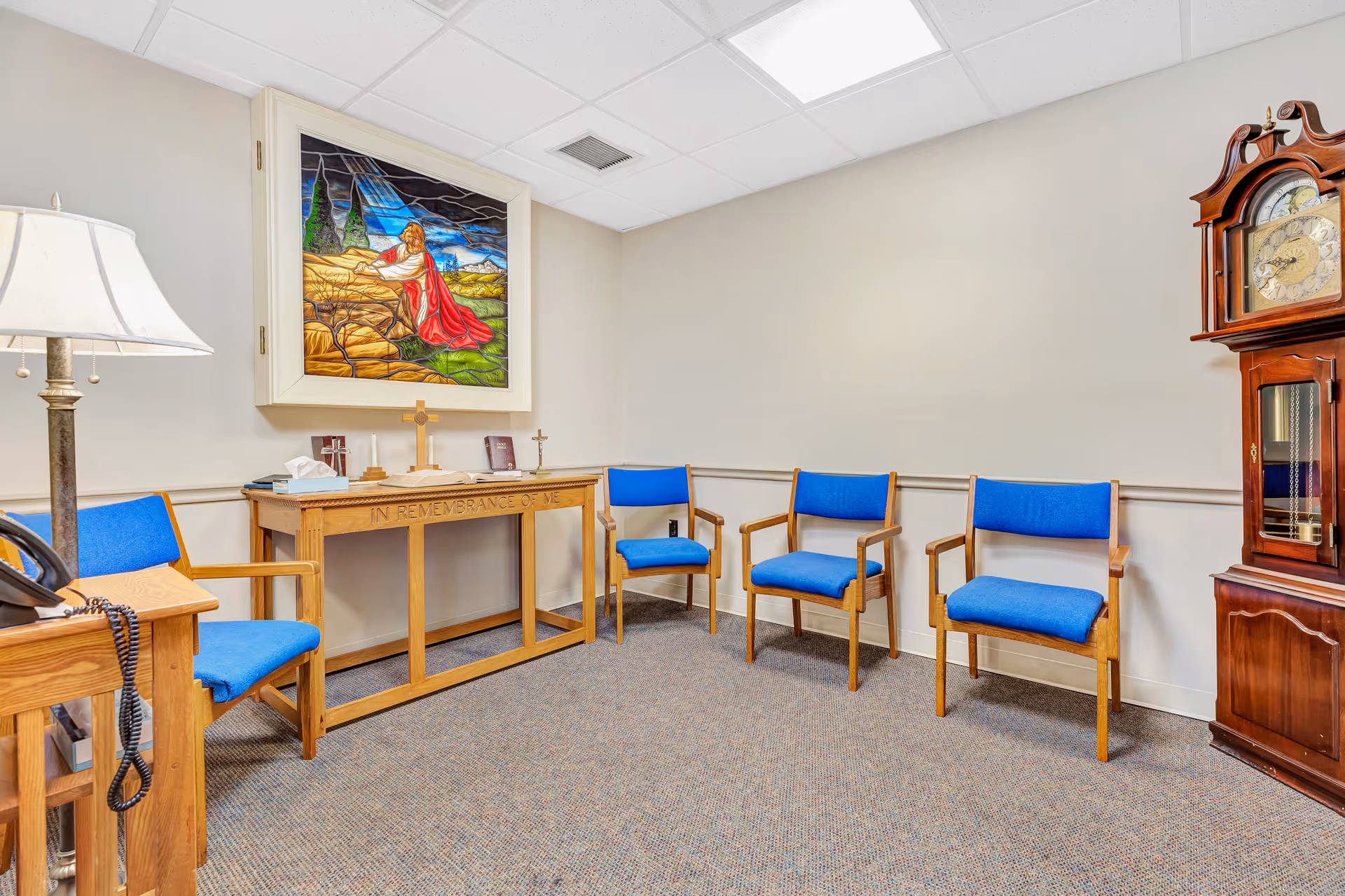 A small room with four wooden chairs with blue cushions arranged against the walls. There is a wooden table with religious items including a cross, a Bible, and a tissue box. Above the table is a framed stained glass artwork depicting a religious scene. A tall wooden grandfather clock stands in the corner, and a floor lamp is visible on the left side.
