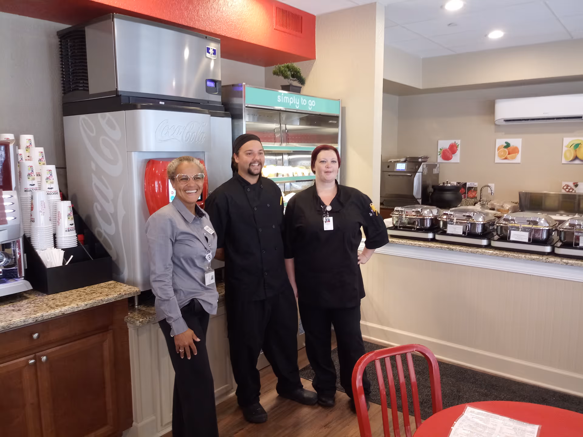 Three staff members standing and smiling inside a dining area with a Coca-Cola beverage dispenser, a refrigerated display case labeled 'simply to go', and a buffet setup with covered food trays on a counter. There are red chairs and a table with a menu in the foreground.