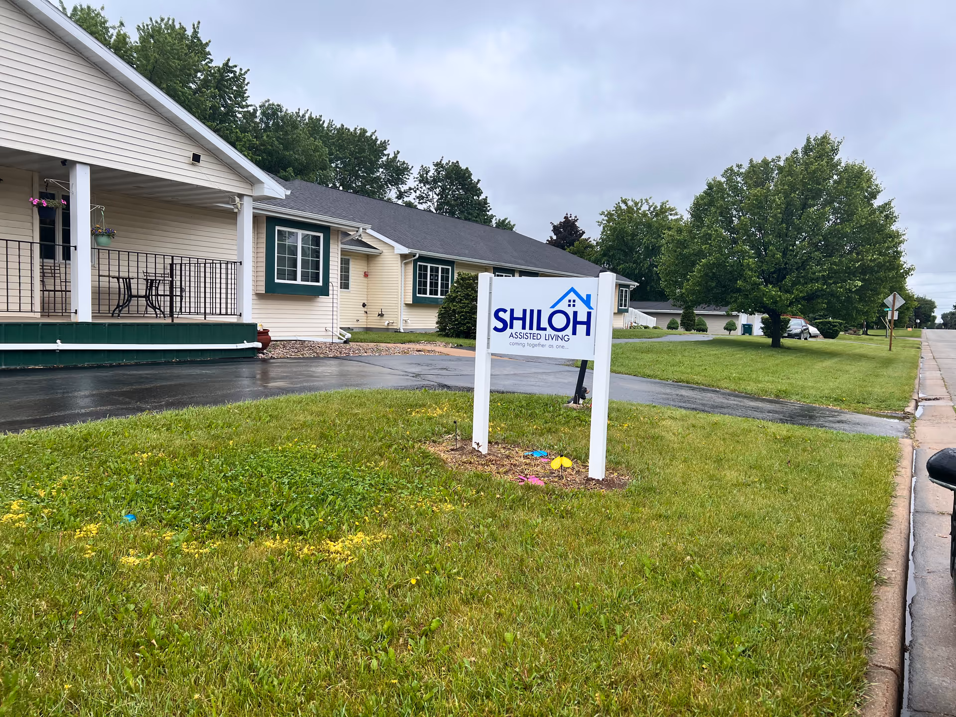 A Shiloh Assisted Living sign on a grassy lawn in front of a single-story beige assisted living building and driveway under a cloudy sky.