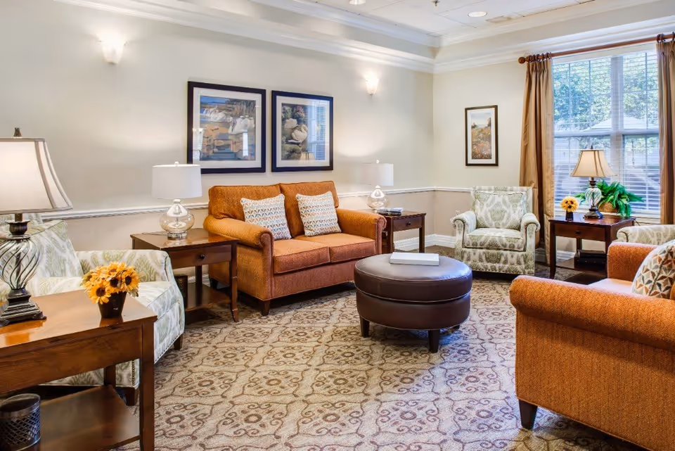 A cozy living room with two orange sofas, two patterned armchairs, a round brown ottoman, wooden side tables with lamps, framed artwork on the walls, and a large window with brown curtains letting in natural light.