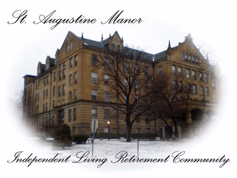 Historic multi-story brick building of St. Augustine Manor with leafless trees and light snow in the foreground.