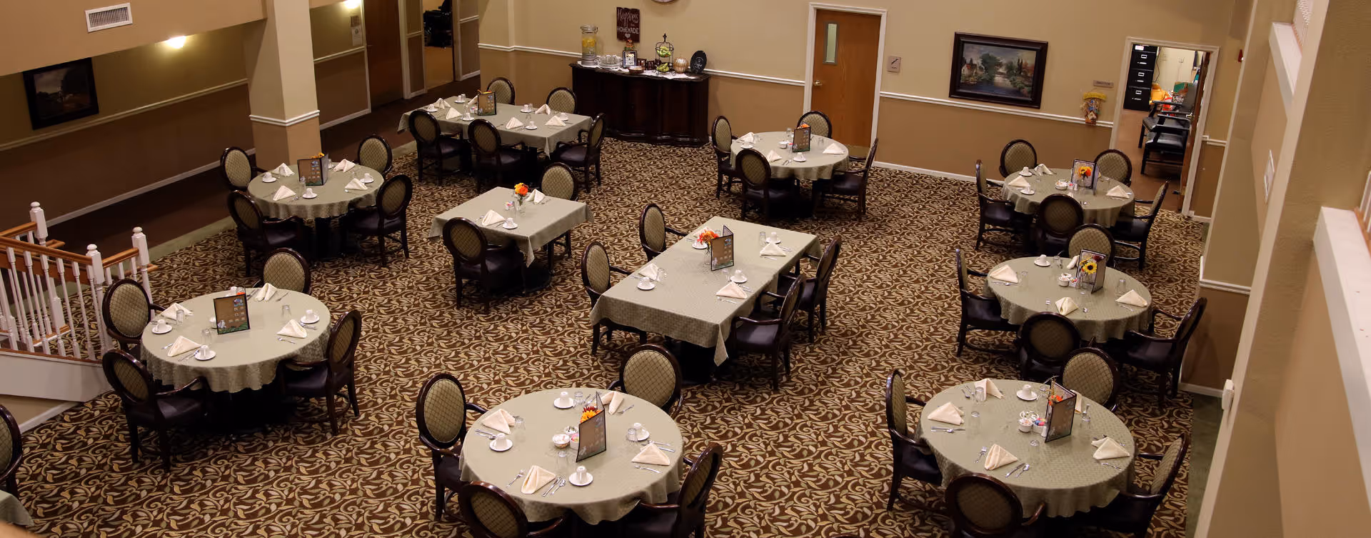 Interior view of a dining room with multiple round and rectangular tables covered with green tablecloths, each set with napkins, plates, and menus. The room has patterned carpet flooring, beige walls, and framed artwork. There is a staircase on the left side and a small buffet area at the back.