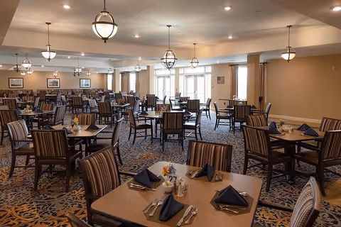 A spacious dining room with multiple tables and chairs arranged neatly. Each table is set with folded navy blue napkins, silverware, and small flower centerpieces. The room is well-lit with hanging pendant lights and large windows allowing natural light to enter. The walls are painted beige, and there is a patterned carpet on the floor. Framed pictures and an American flag are visible on the walls.