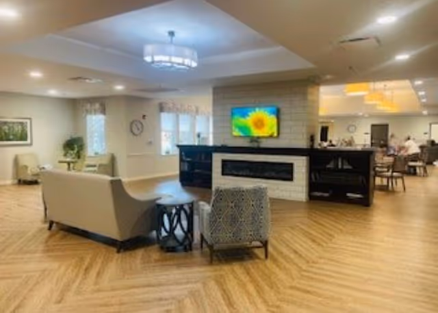 A spacious senior living common area with wood-patterned flooring, a modern fireplace with a TV mounted above it displaying a sunflower image, comfortable seating including a sofa and armchairs, and a dining area visible in the background with people seated at tables. The room is well-lit with ceiling lights and has windows with curtains.