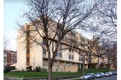Four-story beige brick nursing facility building with bare trees in front, a grassy lawn, and cars parked along the street.