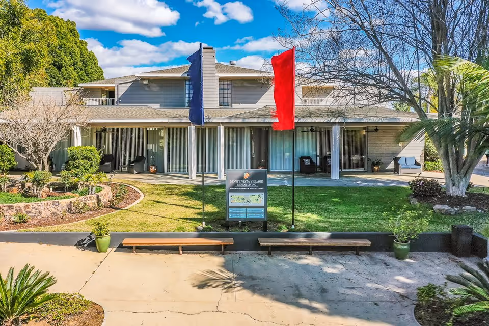 Exterior view of Monte Vista Village senior living facility showing a two-story building with a covered patio area, sliding glass doors, and outdoor seating. In front of the building is a green lawn with two tall flags, one blue and one red, and a sign displaying the facility name and information. The area is landscaped with trees, shrubs, and potted plants under a partly cloudy sky.