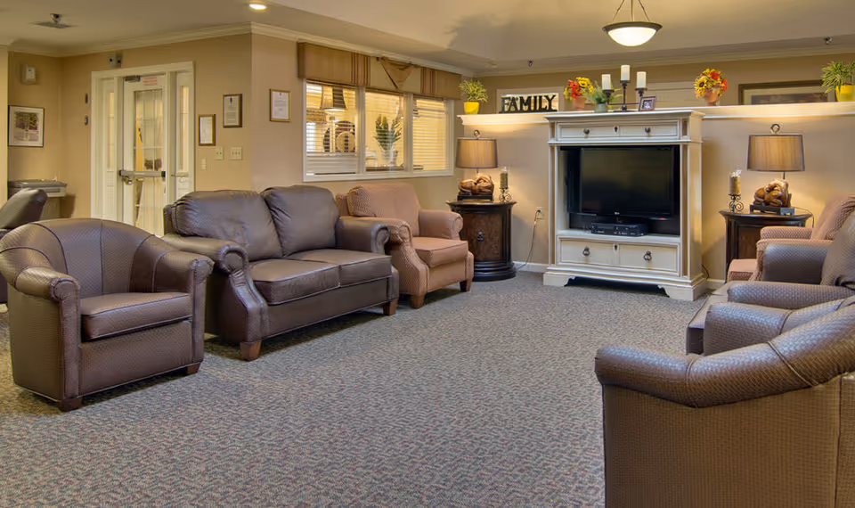 A cozy senior living facility common area with several brown leather armchairs and a loveseat arranged around a white entertainment center with a flat-screen TV. The room has beige walls, carpeted floor, two side tables with lamps, and decorative plants and flowers on shelves above the TV. A window with blinds is visible on the left wall.