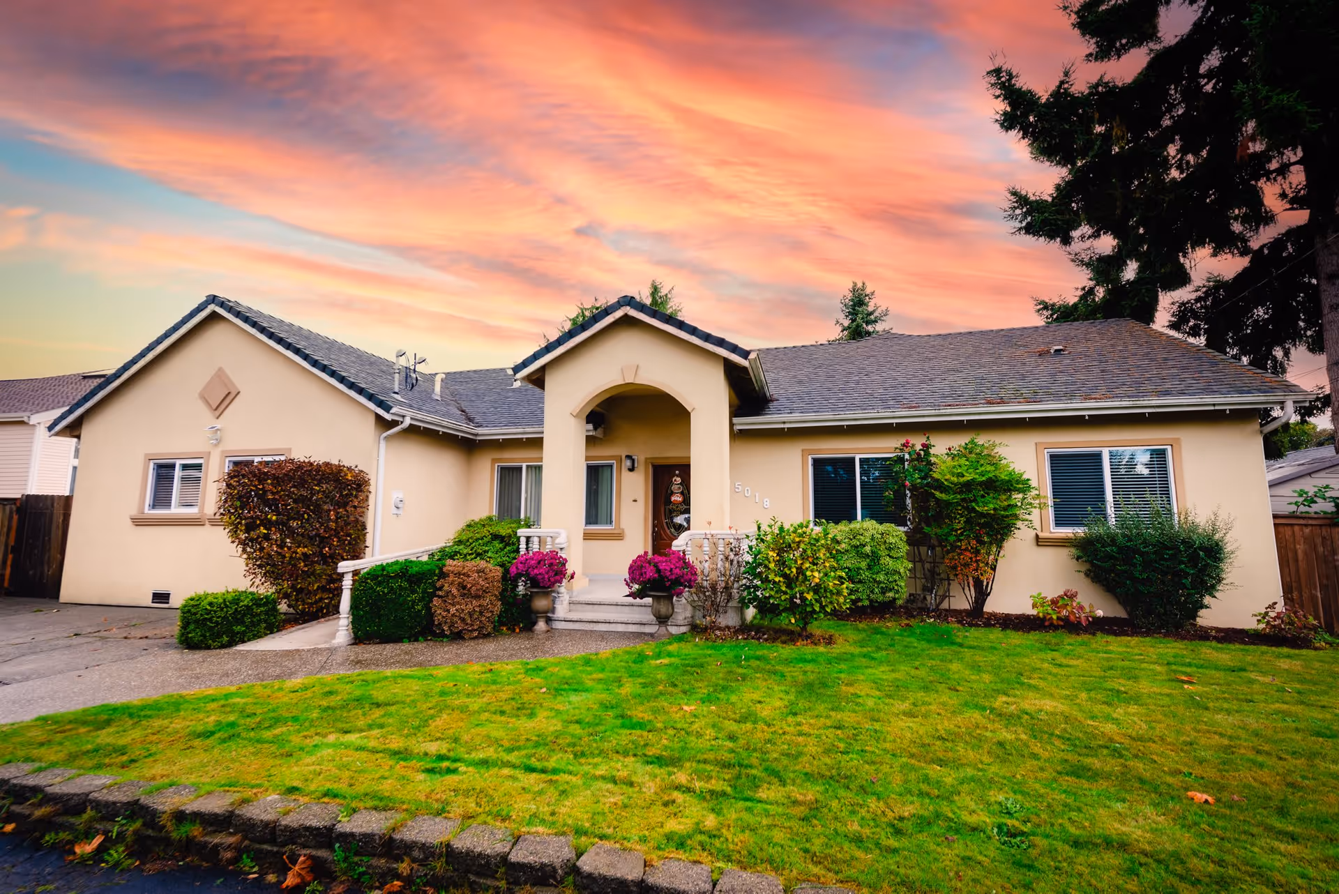 Exterior view of a single-story beige house with a well-maintained green lawn and various bushes and flowers in front. The house has a central entrance with an arched porch and a decorative front door. The sky above shows a colorful sunset with pink and orange clouds.