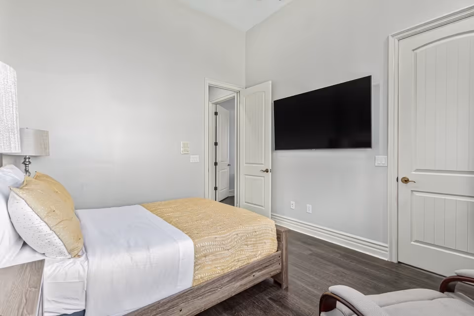 Bedroom with a bed dressed in white and gold bedding, a wall-mounted flat-screen TV, open doorways, and an armchair.