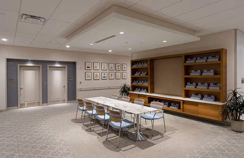 A modern, well-lit interior room with a long rectangular table surrounded by eight chairs with light blue seats. The room has a patterned carpet and neutral-colored walls. On one side, there are two closed doors with signs, and on the adjacent wall, there are framed pictures arranged in a grid. A built-in wooden shelving unit with multiple gray storage bins is on the right side, along with two potted plants.
