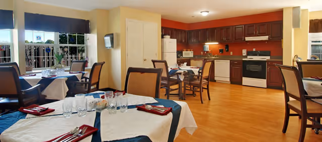 Dining room with several set tables and chairs in the foreground and a full kitchen with appliances in the background.