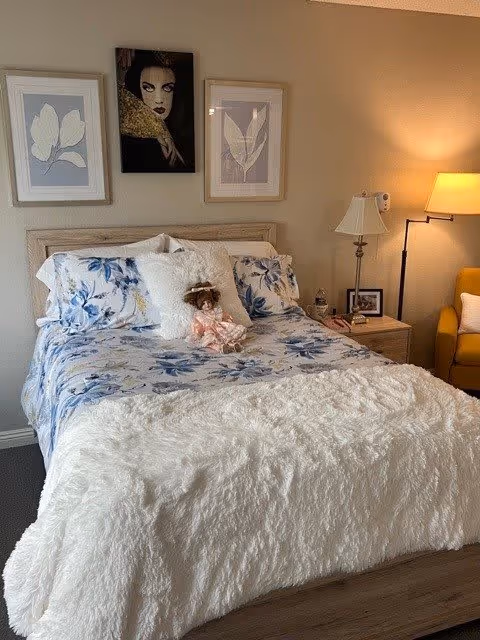 A cozy bedroom with a bed featuring blue and white floral bedding and a fluffy white blanket. A decorative doll is placed on the bed. Above the bed are three framed artworks, two with leaf designs and one portrait of a woman. To the right of the bed is a wooden nightstand with a lamp, a framed photo, and a water bottle. A yellow armchair and a floor lamp are also visible in the corner.