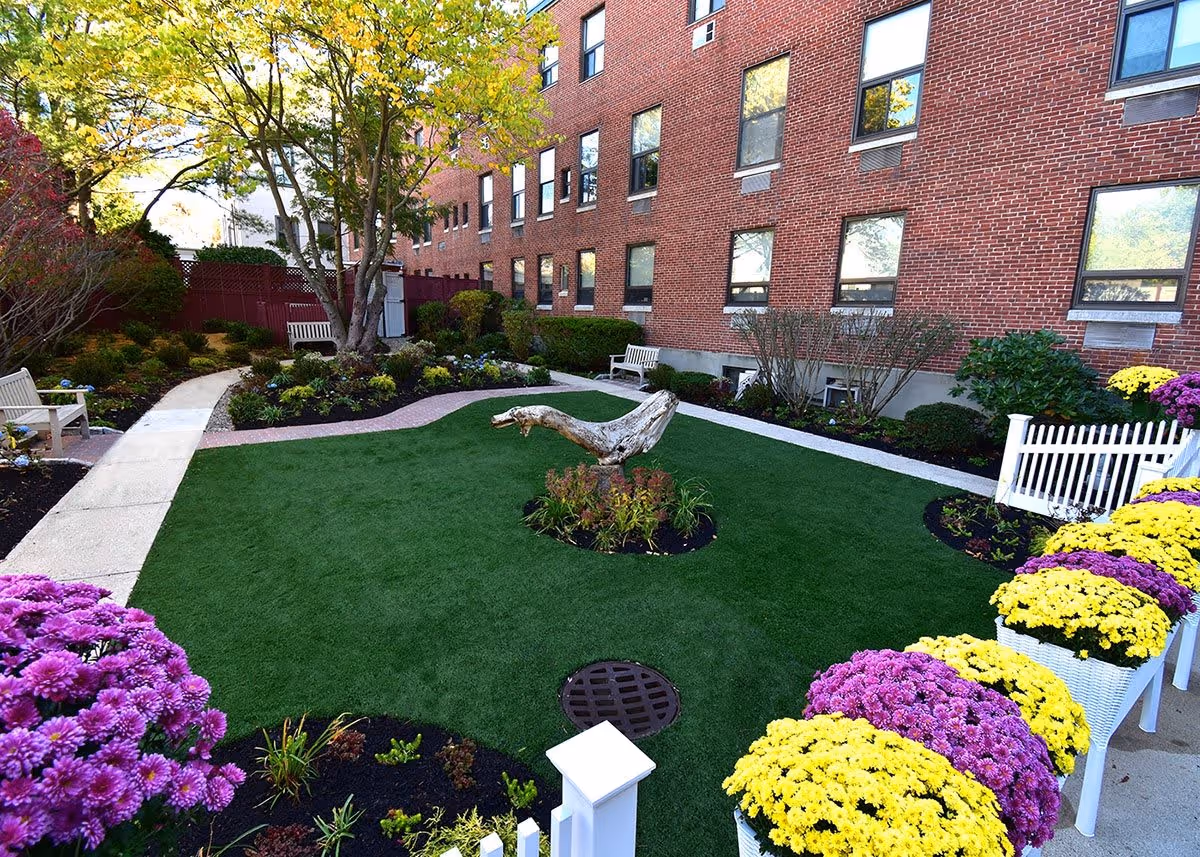 A well-maintained outdoor garden area at a senior living facility with green grass, colorful flower beds featuring purple and yellow flowers, a tree in the center, white benches along the walkway, and a red brick building in the background.