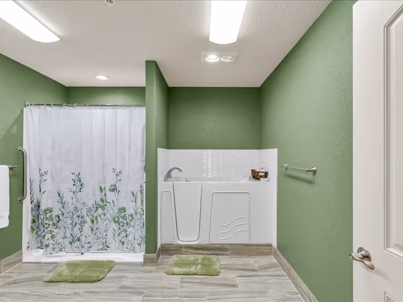 A bathroom with green walls and tiled floor featuring a walk-in bathtub with a faucet and a shower area with a white curtain decorated with green leafy patterns. There are two green bath mats on the floor, a towel hanging on a silver bar, and a door partially visible on the right side.
