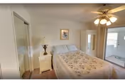 Bright bedroom with a floral-quilted bed, nightstand and lamp, mirrored closet doors, ceiling fan, and a glass-door cabinet leading to an adjacent sunroom.