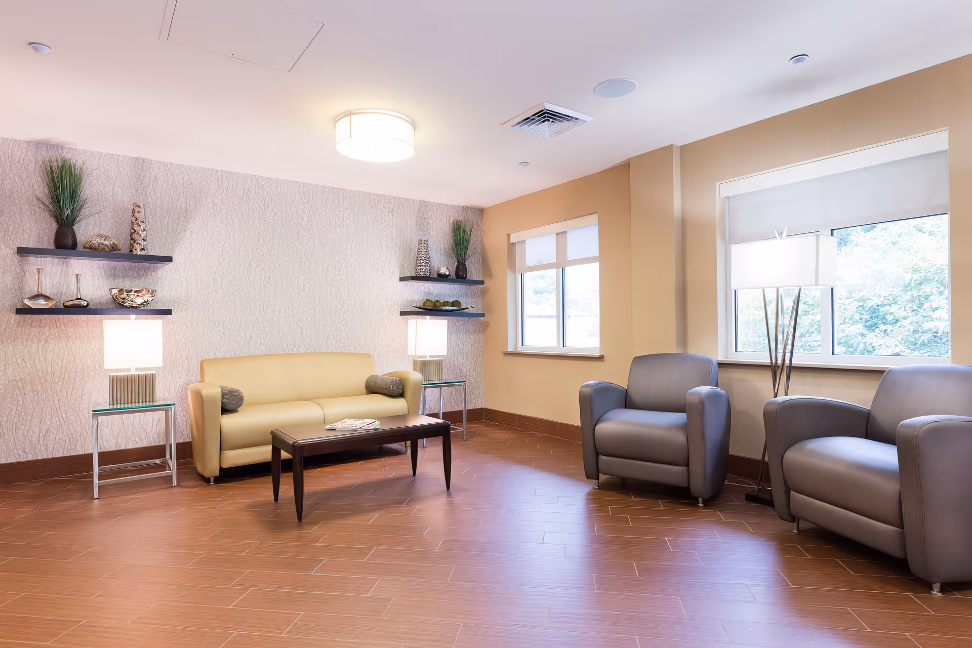 A bright communal living room with a beige sofa, two gray armchairs, a coffee table, wall shelves with decor, and lamps.