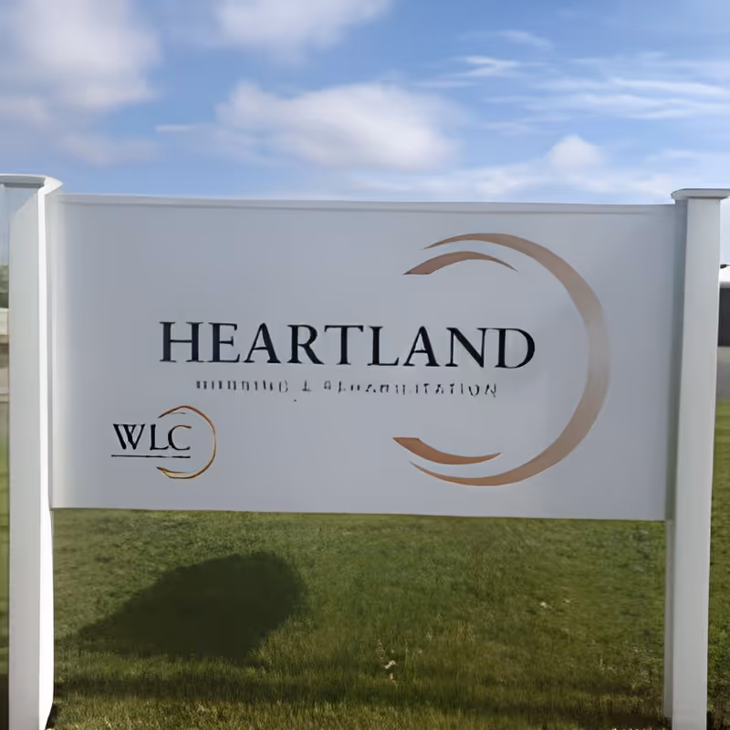 White outdoor sign on a grassy lawn displaying the word "HEARTLAND" with a circular logo and "WLC" marking.