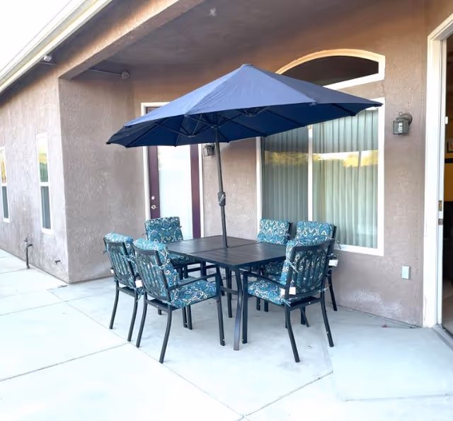 Outdoor patio area with a black metal table and six cushioned chairs with blue patterned fabric. A large blue umbrella is open above the table. The patio is adjacent to a building with beige stucco walls, windows, and a door.
