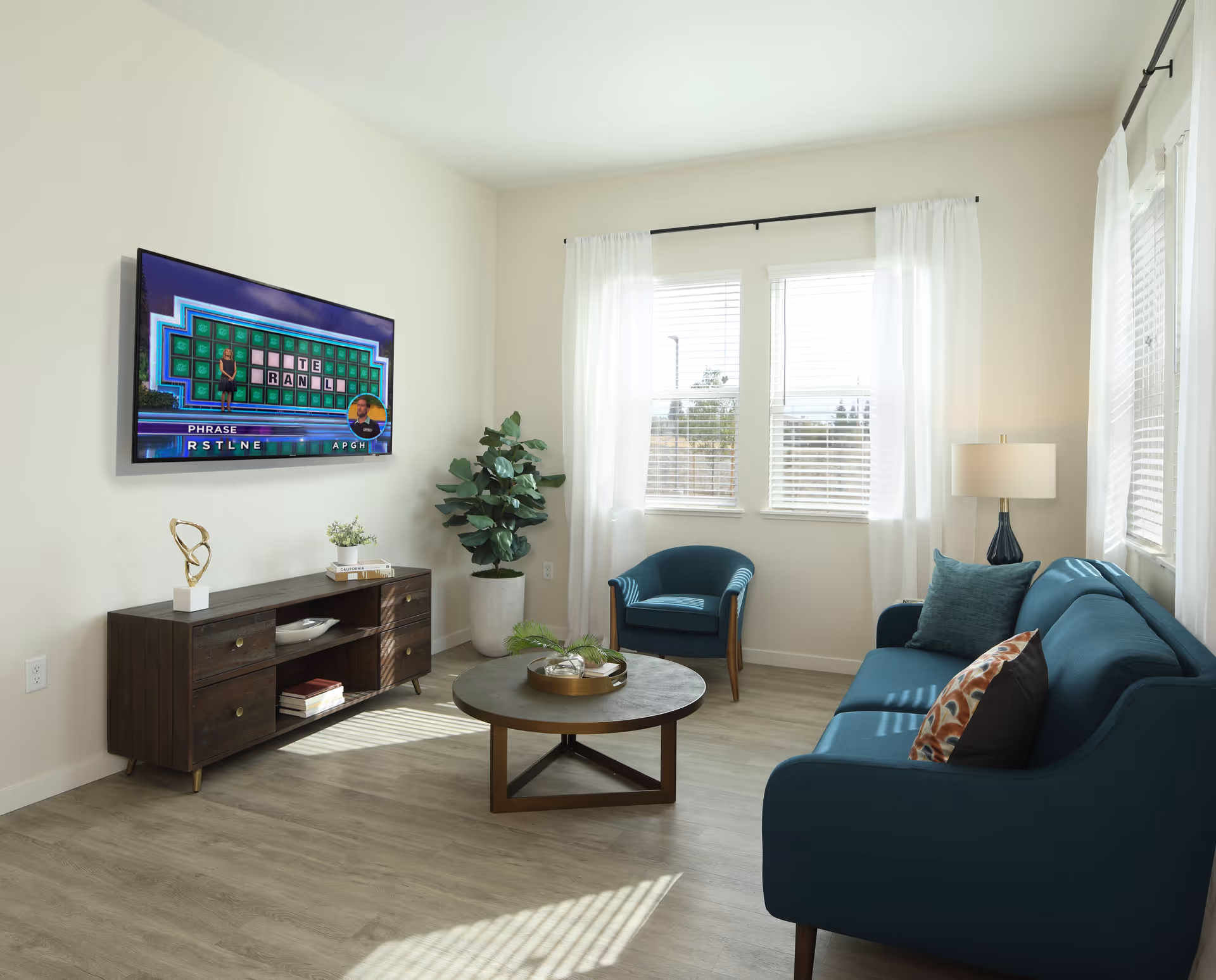 A bright living room with a teal sofa and matching armchair, a round wooden coffee table with a decorative tray, a wooden TV stand with books and decor, a large potted plant, and a wall-mounted flat-screen TV displaying a game show. The room has light-colored walls, wood flooring, and windows with white curtains letting in natural light.