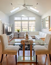Bright living room with vaulted ceiling skylights, large window, white sectional sofa, glass dining table with chairs, TV stand and hardwood floors.